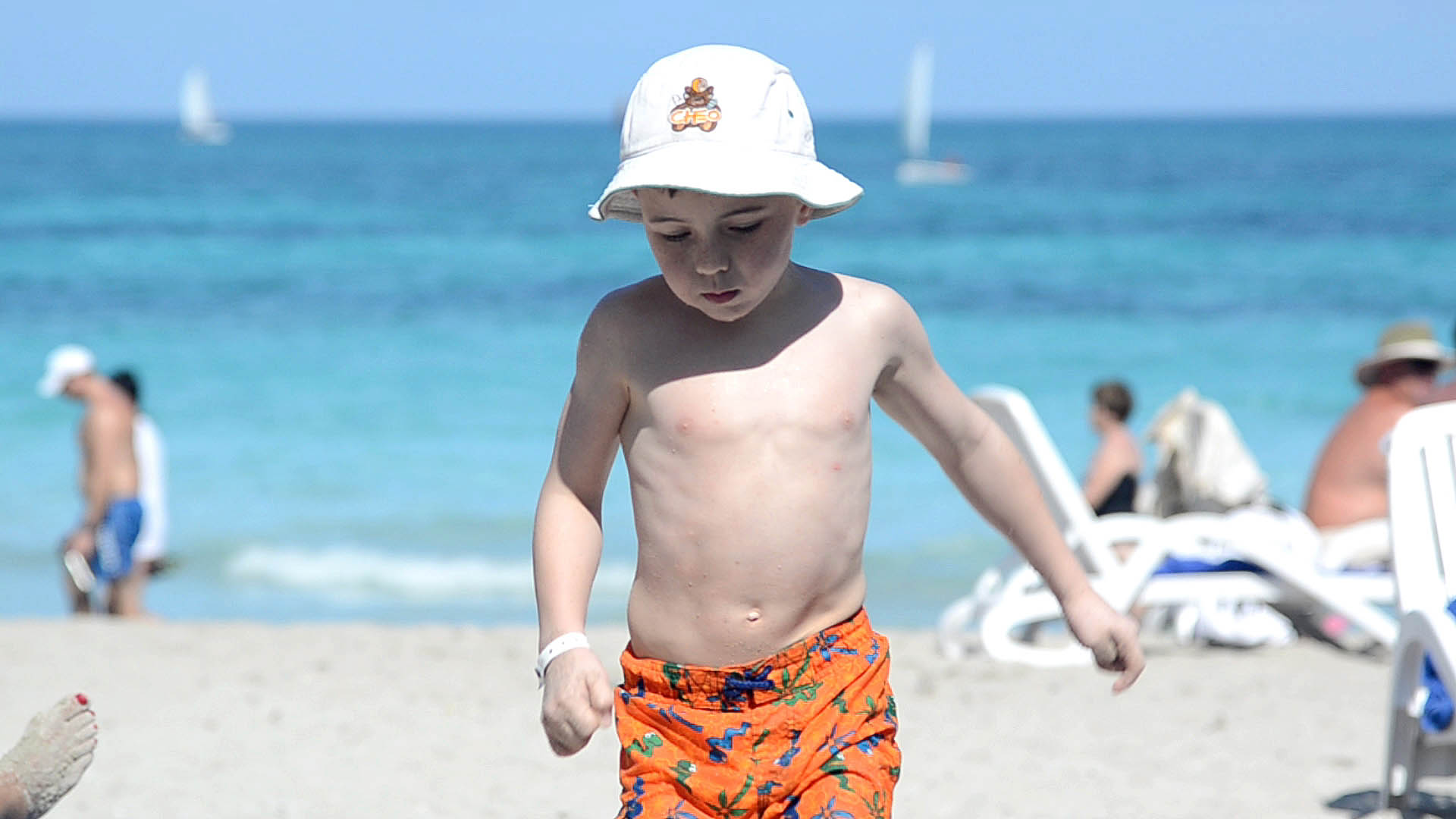 A medium shot of a 7 year old beach boy with bucket hat, running up the beach toward the camera with the beach and the ocean in the background at the Iberostar Laguna Azul Beach Resort in Varadero Cuba