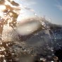The photograph shows a somewhat backlit wake surfer surfing towards the back of the boat about to hop onto the back of the boat. There is a spash of water over the GoPro camera lens in the forground and the wake wave of the boat in the background The wake surfer is wear a half wetsuit.
