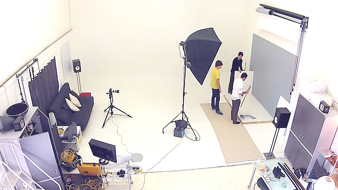 The photograph shows photographers, assisstants and stylist setting up photography gear such as cameras and lighting for a photo shoot.
