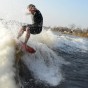 The photograph shows a wake surfer wake surfing in a wetsuit high on the wake wave with cold water spashing all around him. The wake surfer has his arms up and his finger is pointing, through the splash of the wave, towards the camera in the boat.