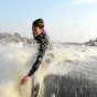 The photograph shows a wake surfer wake surfing in a wetsuit high on the wake wave with cold water spashing all around him. The wake surfer caught the front of his wake surfing board in the wake wave and is falling into the wake wave as the spray flies up into his face