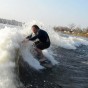 The photograph shows a wake surfer wake surfing in a wetsuit high on the wake wave with cold water spashing all around him. The wake surfer is crouched one his wake surf board with his arms in front of him catching the wake wave causing spray to fly up into his face as he surfs towards the camera in the boat.