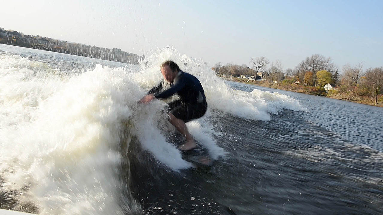 The photograph shows a wake surfer wake surfing in a wetsuit high on the wake wave with cold water spashing all around him. The wake surfer is crouched one his wake surf board with his arms in front of him catching the wake wave causing spray to fly up into his face as he surfs towards the camera in the boat.