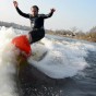 The photograph shows a wake surfer wake surfing in a wetsuit high on the wake wave with cold water spashing all around him. The wake surfer has his arms up as he surfs high on the wake wake towards the camera in the boat.