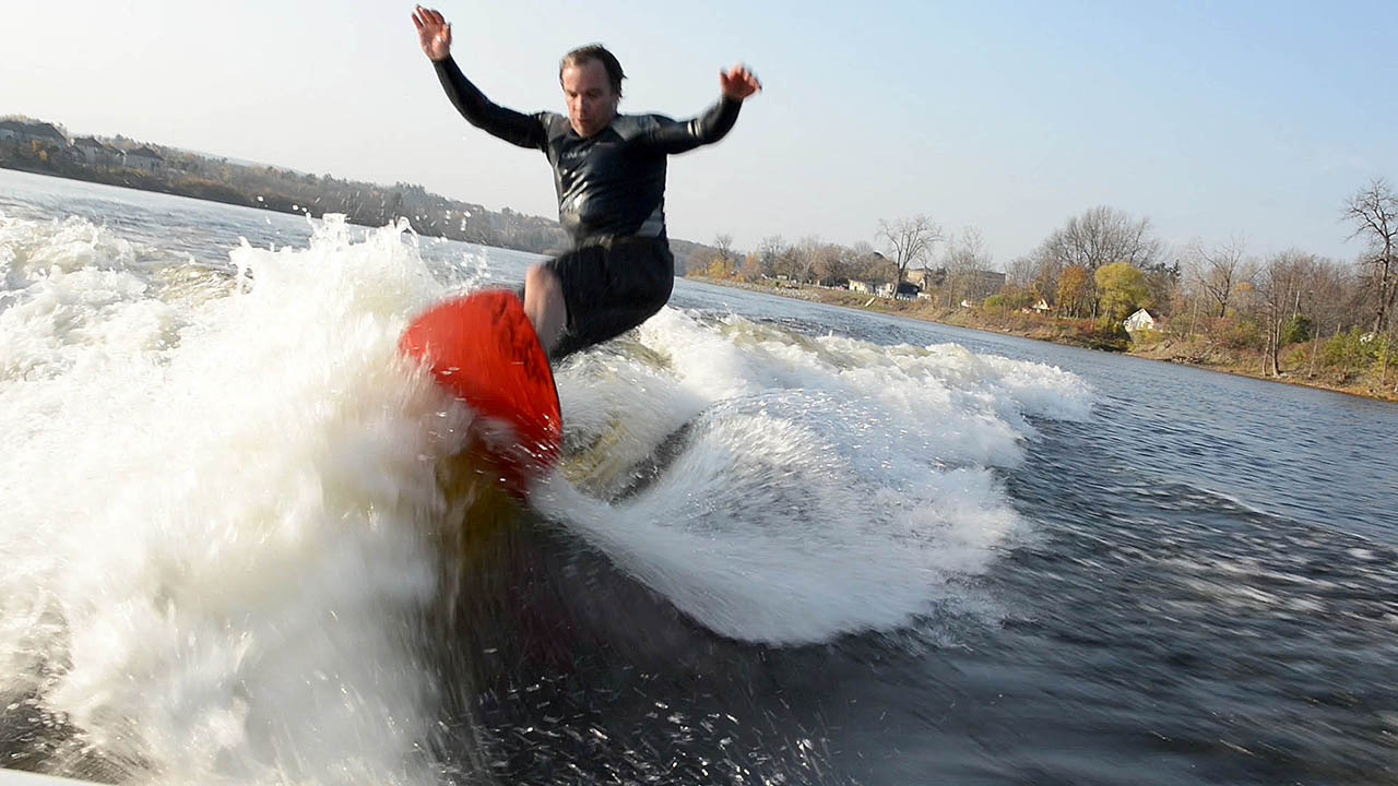 The photograph shows a wake surfer wake surfing in a wetsuit high on the wake wave with cold water spashing all around him. The wake surfer has his arms up as he surfs high on the wake wake towards the camera in the boat.