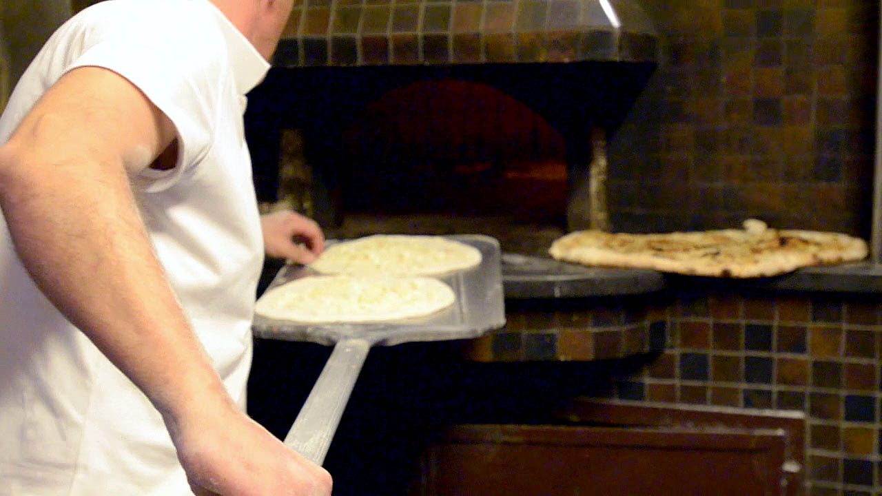 A pizzaiolo checking the pizza dough as he puts the pizza in the wood fire oven