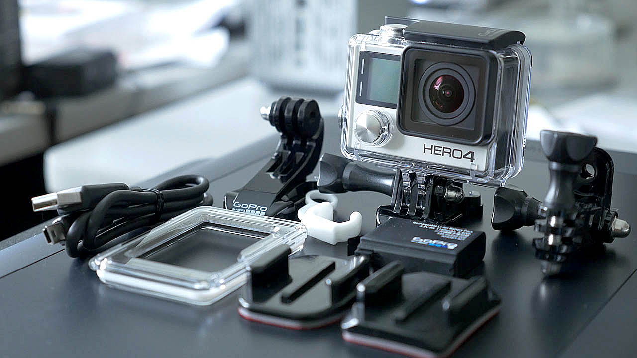 The photograph shows the GoPro Hero 4 Black Edition on a bit of an angle so the side and front are visible with the Hero 4 logo very prominent on the front of the GoPro and all the accessories that come with the GoPro Hero 4 Black layed out infront ot the GoPro.