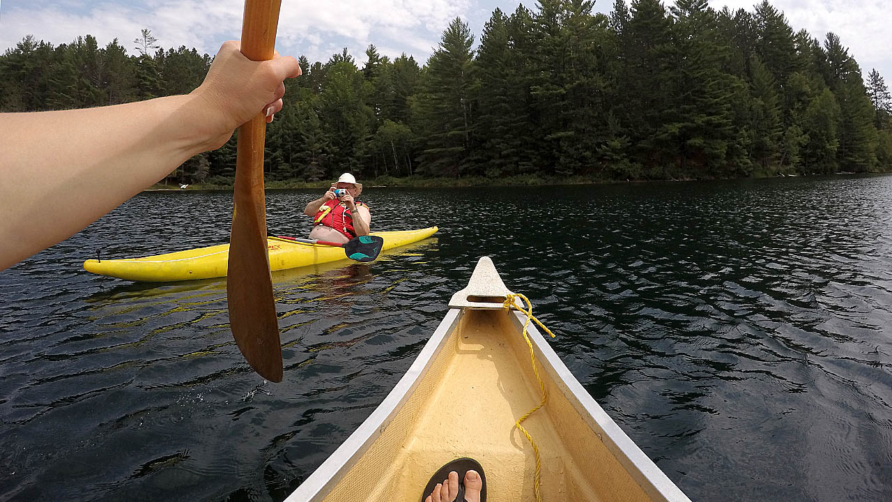 The photograph is point of view photograph from a GoPro Hero 4 Black camera strapped to the life jacked of a paddler in the front of the canoe with one hand and with a paddle and the bow of the canoe visible in the forground and the water of the lake, the shoreline and a kayaker visible in the background
