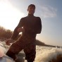The photograph shows a somewhat backlit wake surfer standing on the back of the boat ajusting the arm of his wet suit with the wake surfboard on the boat beside him. The wake surfer is wear a full wetsuit.