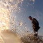 The photograph shows a very low angle view of a wake surfer standing staight up on the wake surfing board with the sun and sky in the background. The wake surfer is wear a full wetsuit and splash is over most of the GoPro camera lens