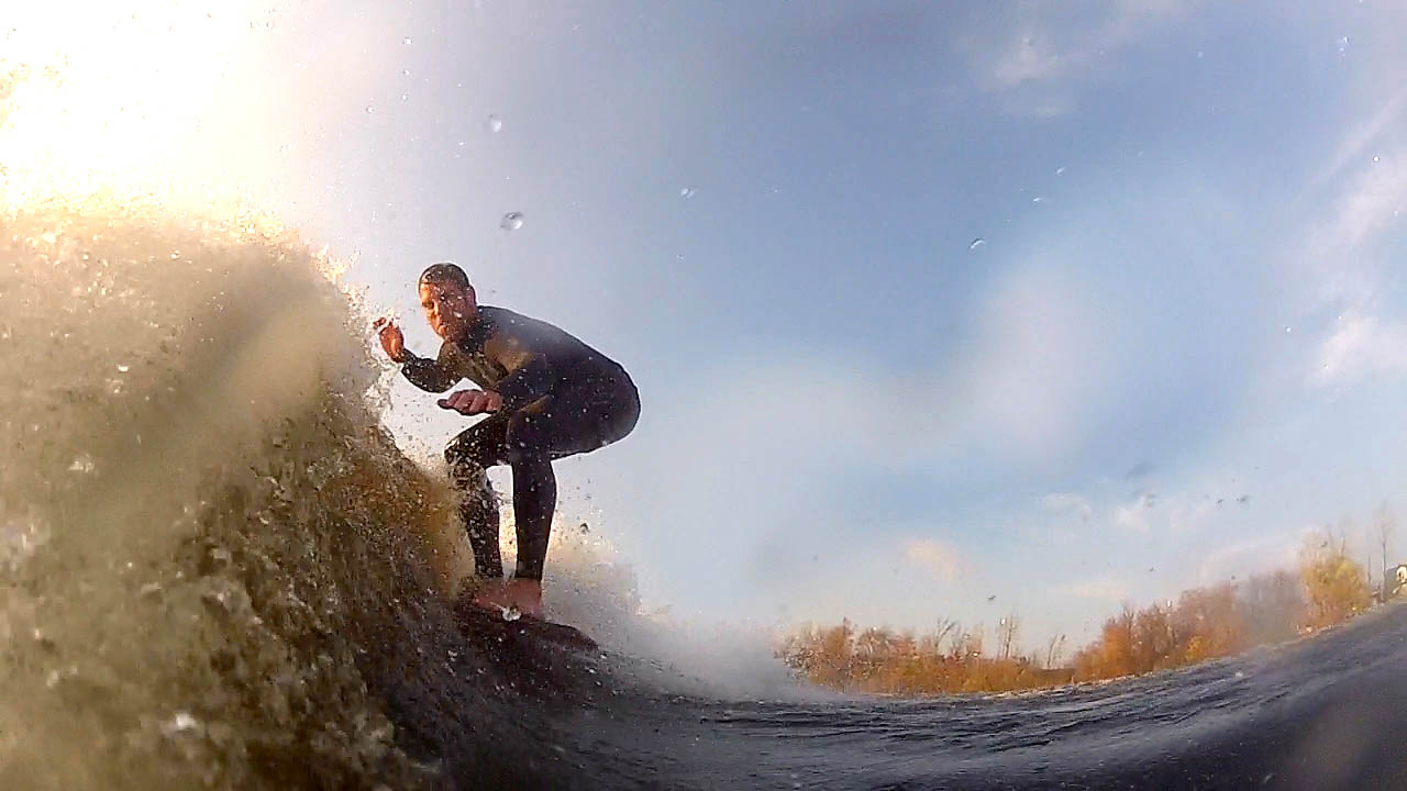 The photograph shows a very low angle view of a wake surfer riding the wake wave towards the back of the boad with the sun and sky and some distant trees in the background The wake surfer is wear a full wetsuit and there is some wake wave splash across the GoPro camera lens