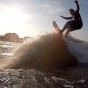 The photograph shows a somewhat backlit wake surfer high on the wake wave with the sun in the background and the back of the wake boat on the left side. The wake surfer is wear a half wetsuit.