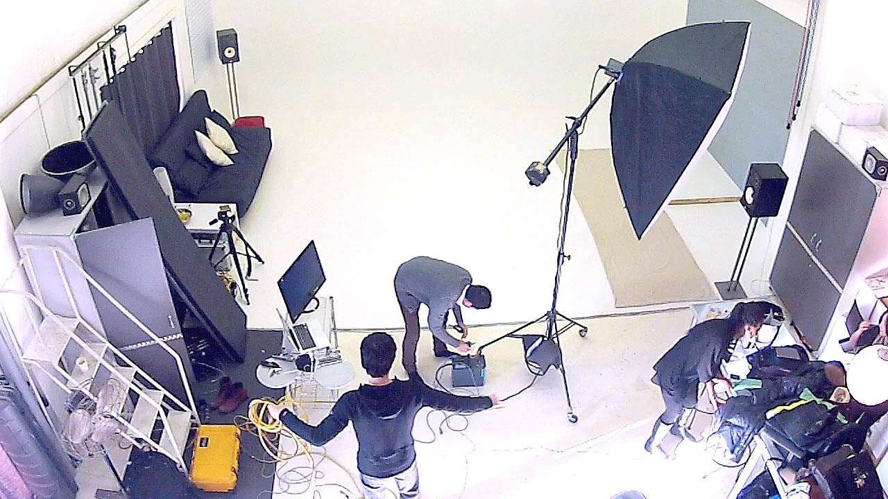 The photograph shows photographers, assisstants and stylist setting up photography gear such as cameras and lighting for a photo shoot.