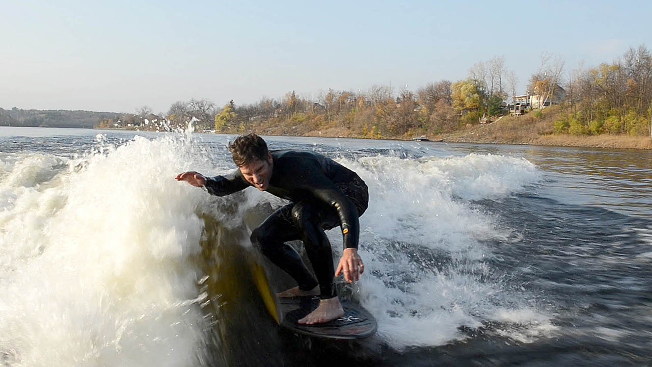 The photograph shows a wake surfer wake surfing in a wetsuit high on the wake wave with cold water spashing all around him. The wake surfer is crouched on the wake surfing board with on hand up catching the spray from the wake wave as he surfs towards the camera in the boat.